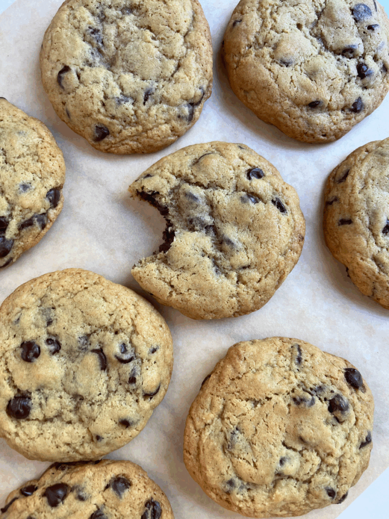 Close-up of freshly baked sourdough chocolate chip cookies on parchment paper, with one cookie showing a bite taken out to reveal the soft, gooey center